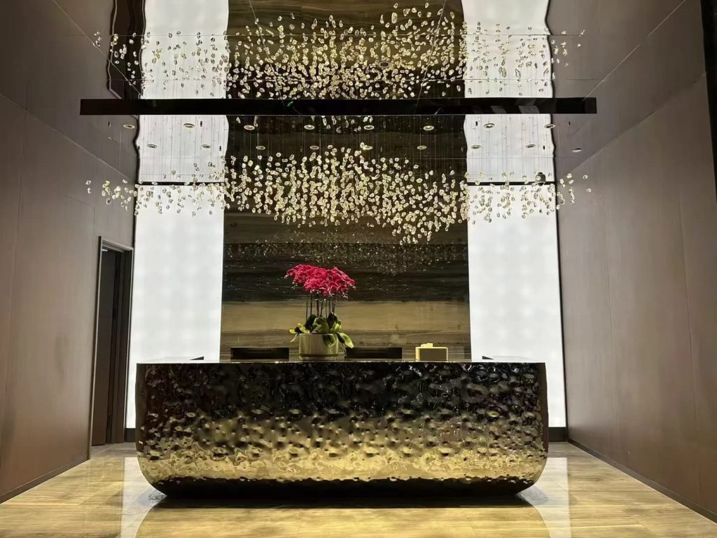 Custom stainless steel art reception desk with flowing water-ripple textured surface, sculpted organic curves, and integrated countertop, displayed in a contemporary lobby.