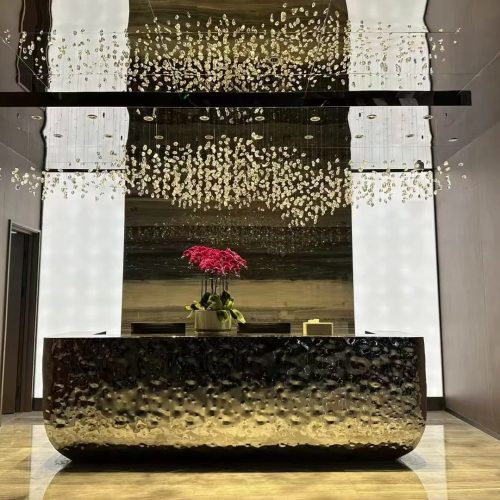 Custom stainless steel art reception desk with flowing water-ripple textured surface, sculpted organic curves, and integrated countertop, displayed in a contemporary lobby.